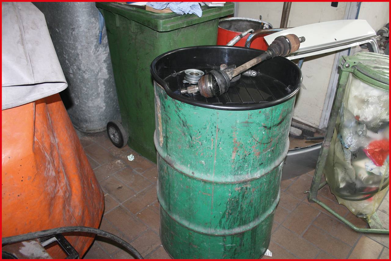 Zielony metalowy beczka stoi w warsztacie. Leży na niej wał napędowy samochodu. W tle widoczne są śmietniki i różne przedmioty warsztatowe.