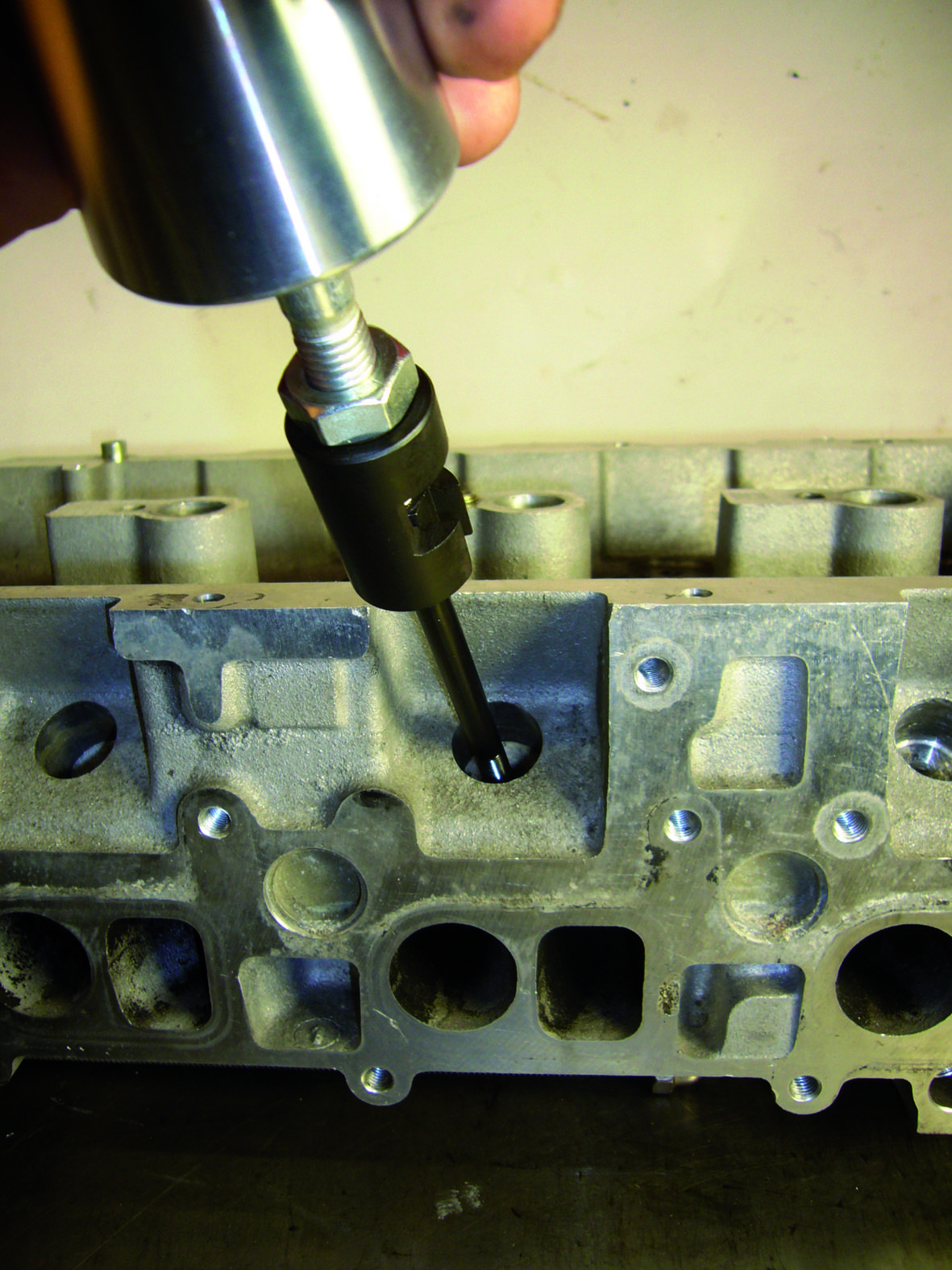 Urządzenie metalowe jest wmontowywane w głowicę cylindra. W tle widoczne są szczegóły głowicy cylindra.