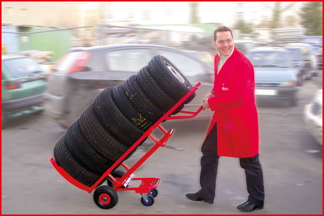 Mężczyzna w czerwonym płaszczu przesuwa wózek z wieloma ułożonymi na stosie oponami samochodowymi na parkingu.
