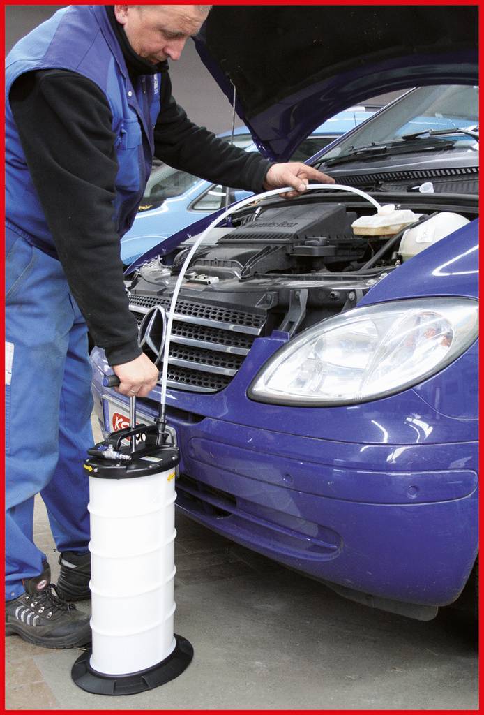 Mechanik używa pompy próżniowej do odsysania płynów z niebieskiego samochodu. Maska silnika jest otwarta.