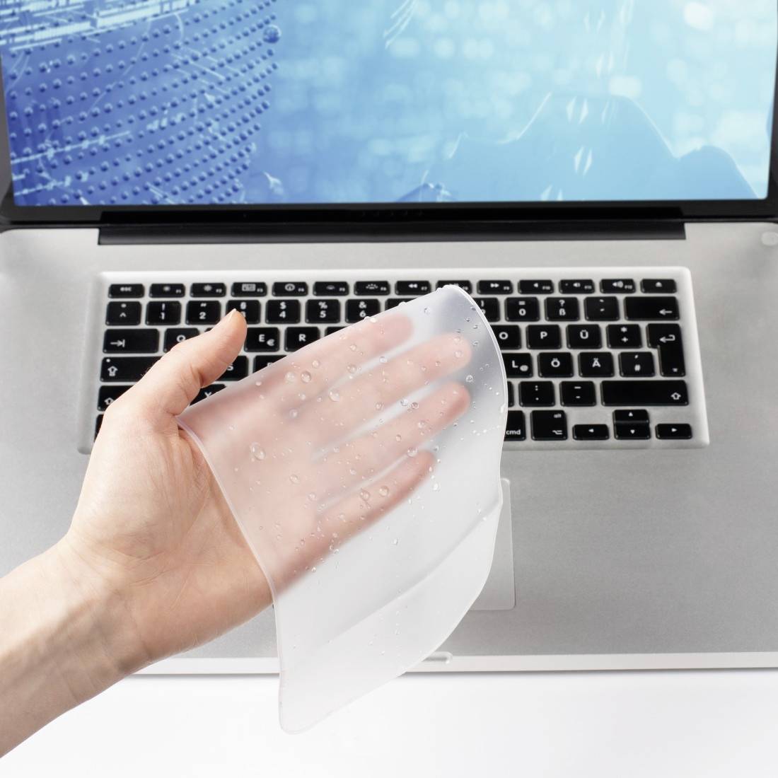 Osoba trzyma przezroczysty, elastyczny pokrowiec silikonowy nad klawiaturą laptopa. Na ekranie laptopa widać abstrakcyjny wzór.