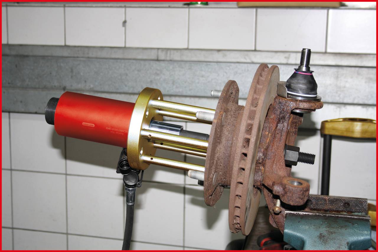 Narzędzie pokazuje demontaż przegubu kulowego na wsporniku koła pojazdu. Przegub kulowy jest częściowo zardzewiały i zamocowany.