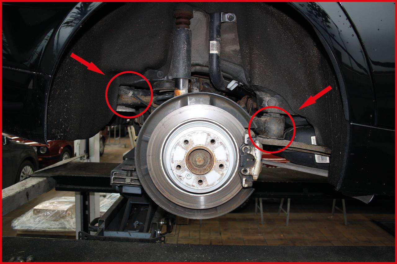 Samochód jest podniesiony, koło zostało zdjęte. Fokus skierowany jest na zawieszenie i elementy osi, z dwoma czerwonymi strzałkami i kołami oznaczającymi konkretne części.