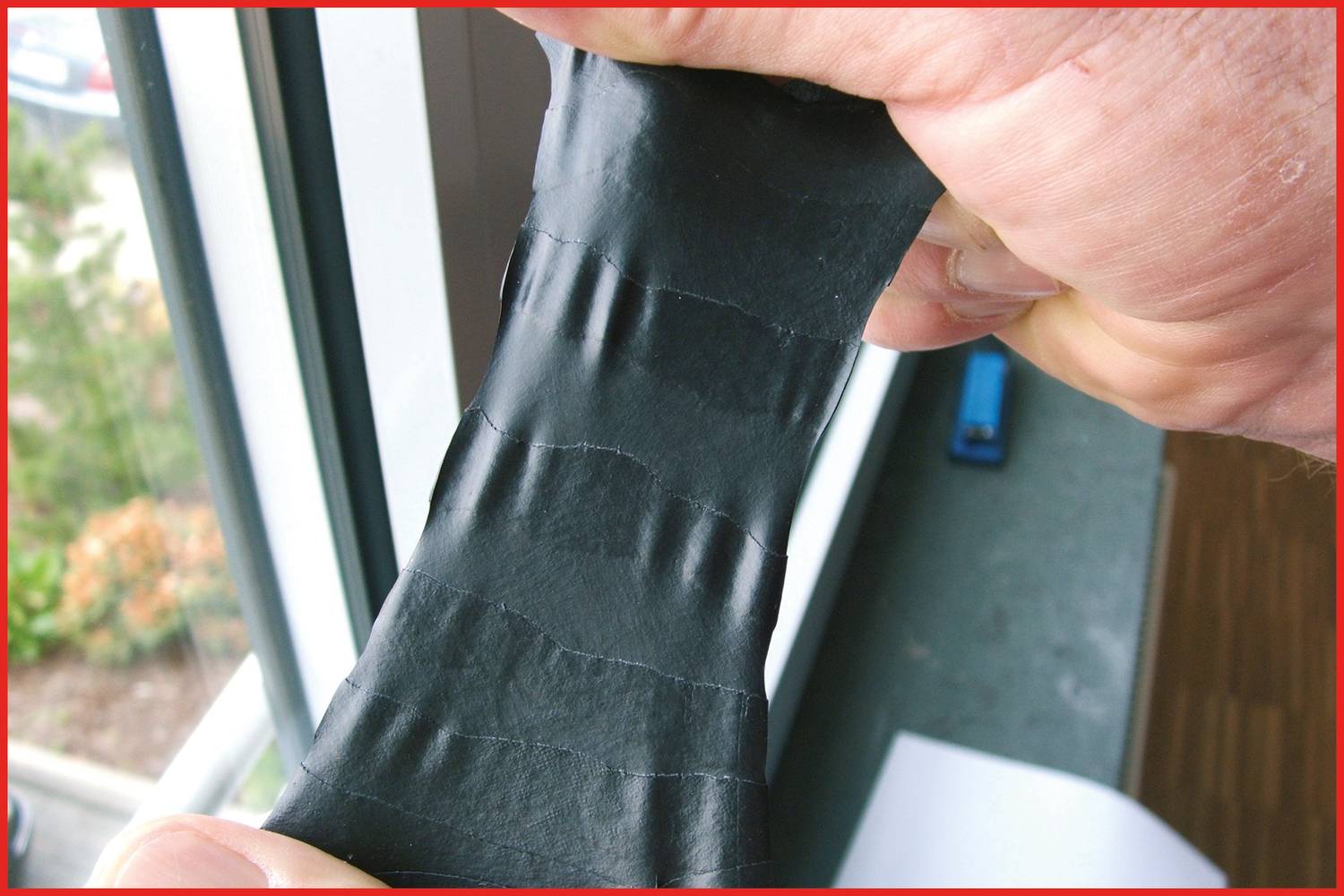 Ręka rozciąga czarny pas materiału. W tle widoczne są okno i fragment dywanu.