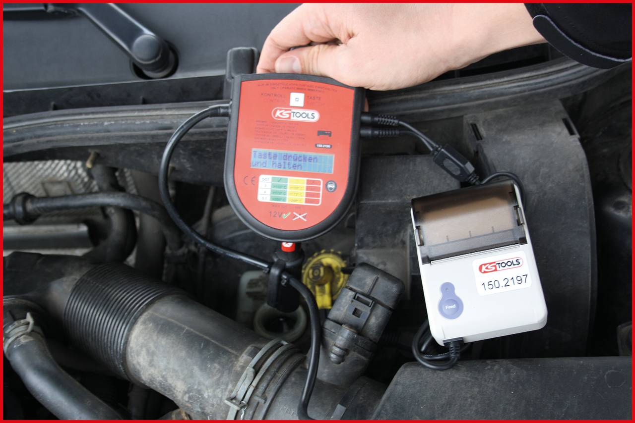 Ręka trzyma narzędzie diagnostyczne z wyświetlaczem, podłączone do części samochodowej. Urządzenie drukujące wyświetla logo 'KS Tools'.