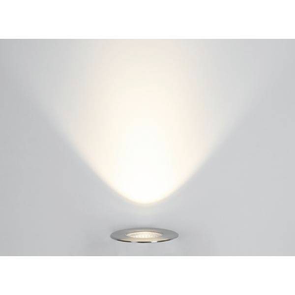 Okrągła, wpuszczona w sufit lampa rzuca ciepłe światło w kształcie stożka, odbijające się od białej ściany.