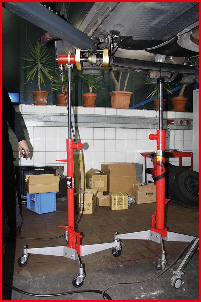 Czerwony podnośnik samochodowy unosi część samochodu w warsztacie. W tle widoczne są doniczki z roślinami i kartony.