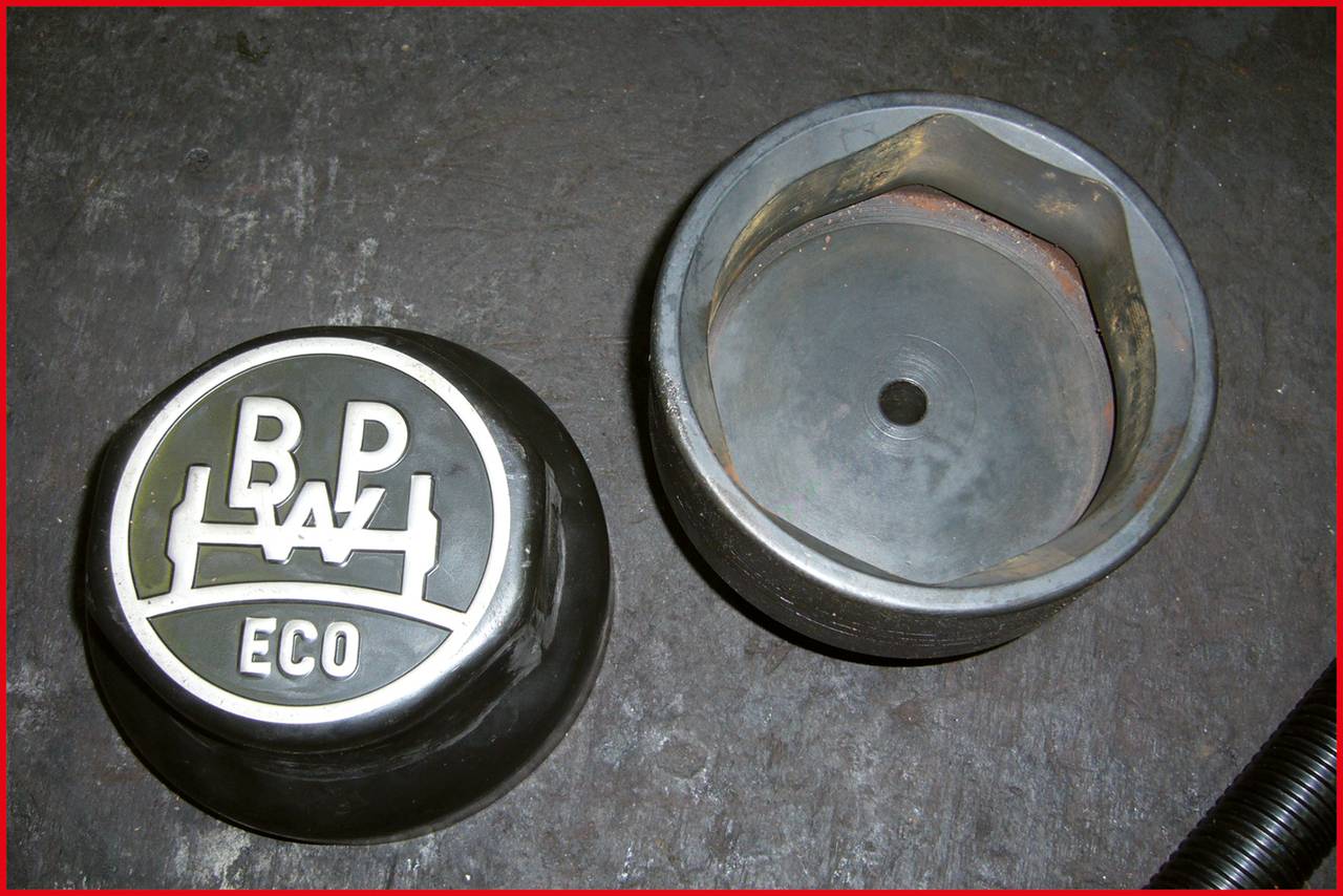 Dwie metalowe komponenty na betonowej podłodze. Po lewej stronie pokrywa z logo 'BP ECO'; po prawej stronie odpowiednie mocowanie bez logo.