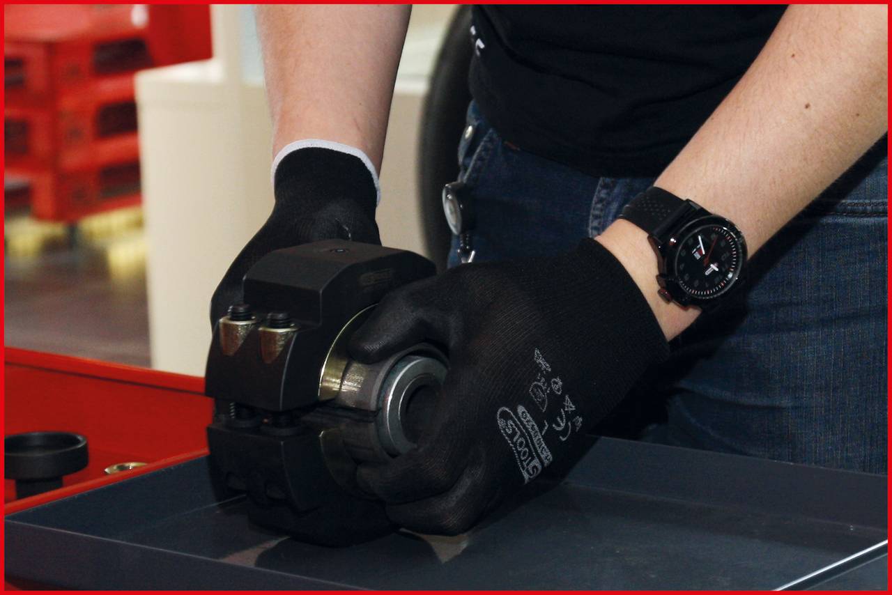 Osoba w czarnych rękawicach pracuje przy części samochodowej w warsztacie. Na tle widoczne są narzędzia.