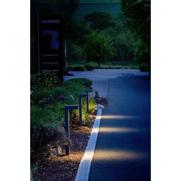 Ulica o zmierzchu z oświetlonymi latarniami wzdłuż chodnika. Lasy i trawy w tle.