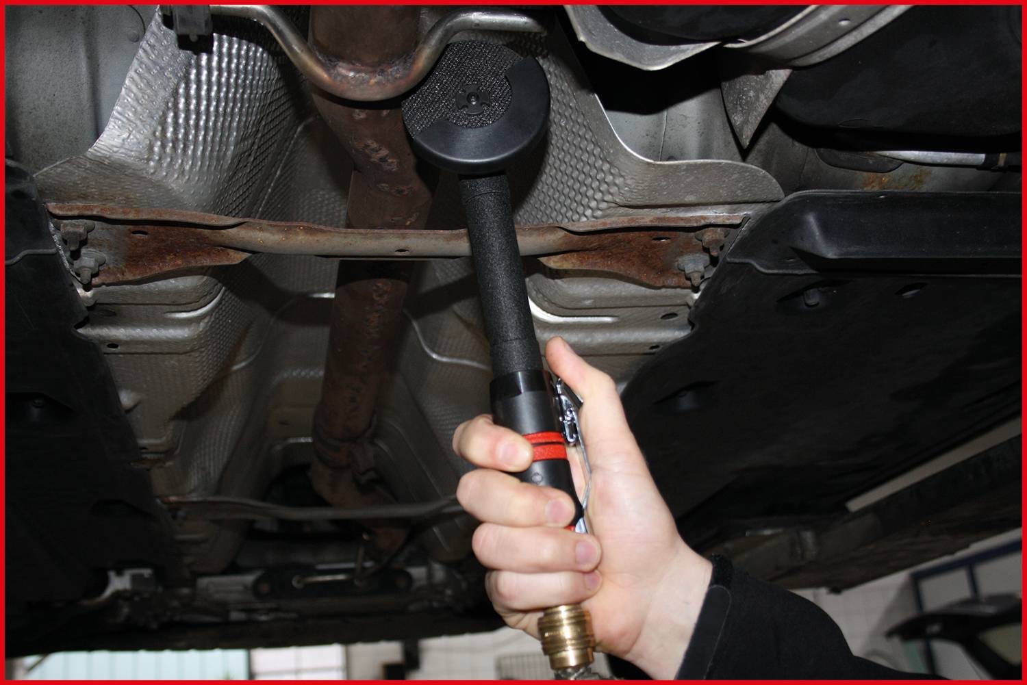 Osoba trzyma miernik emisji spalin przy wydechu pojazdu, aby zmierzyć poziom emisji. Samochód znajduje się na podnośniku warsztatowym.