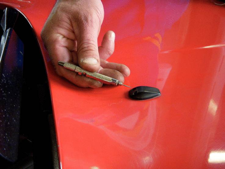 Dłoń trzyma małe metalowe narzędzie obok czarnego lusterka bocznego na błyszczącej czerwonej drzwi samochodu.
