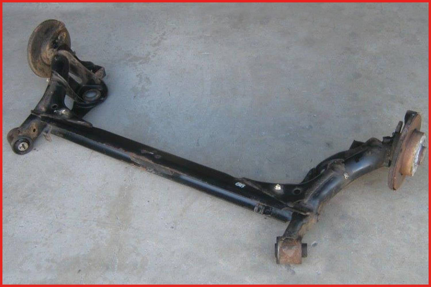 Czarna tylna oś pojazdu leży na betonowej podłodze. Widoczne są dwie piasta kół na końcach oraz poprzeczne belki wzmacniające.