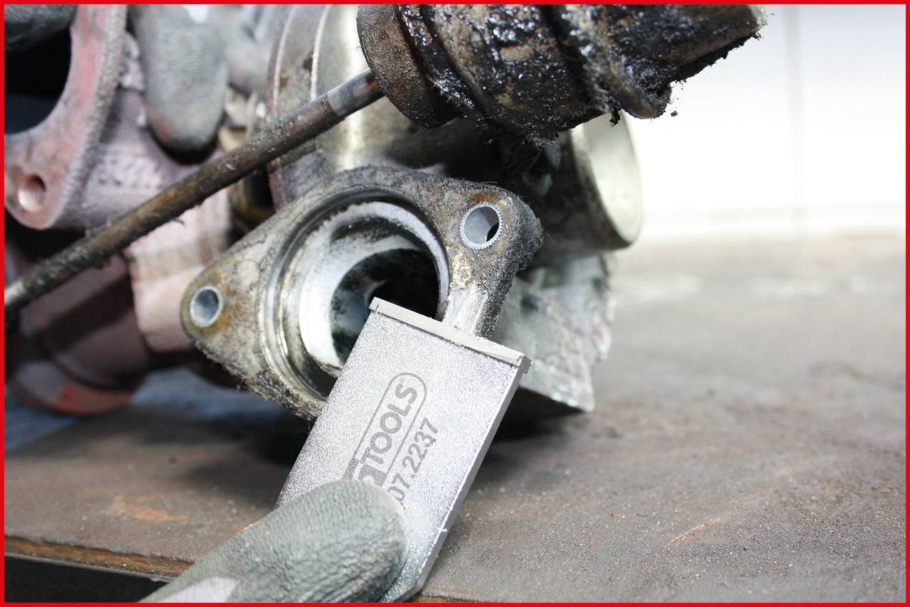 Na stole warsztatowym leży zardzewiała część samochodowa, a narzędzie z napisem 'TOOLS' jest używane do usuwania nawarstwień.