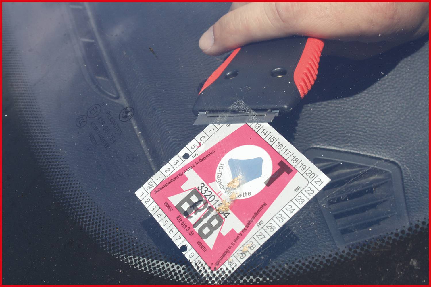 Ręka używa skrobaczki, aby usunąć winietę z przedniej szyby. Winieta ma czerwono-białe wzornictwo z czarnym tekstem.