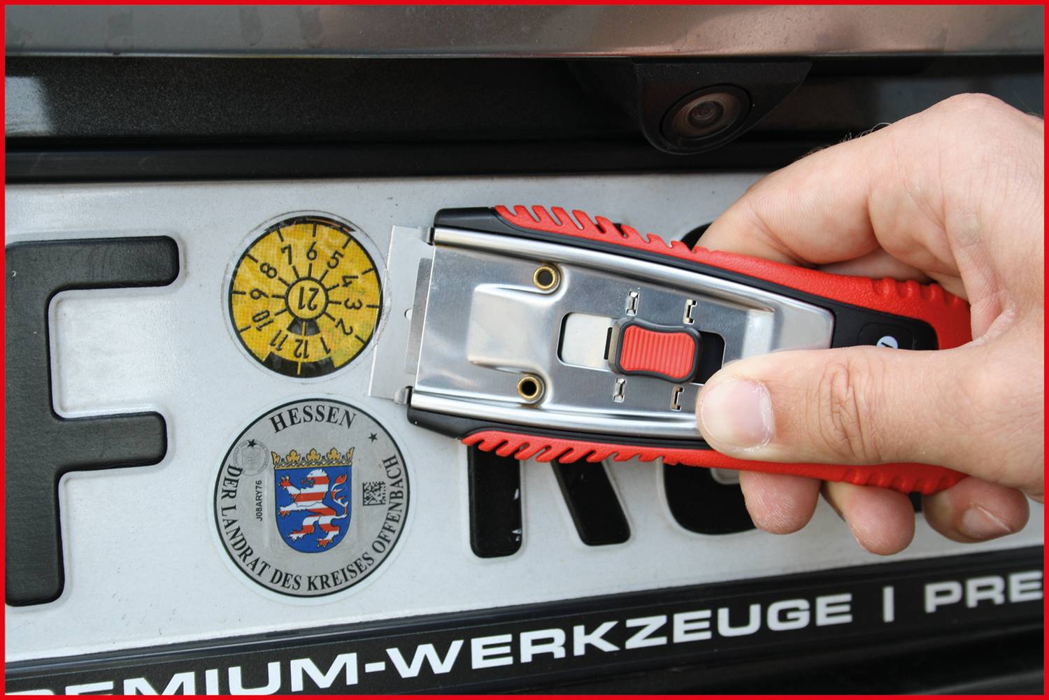 Jedna ręka trzyma nóż do tapet nad tablicą rejestracyjną z naklejką kontrolną i etykietą powiatu Offenbach w Hesji.