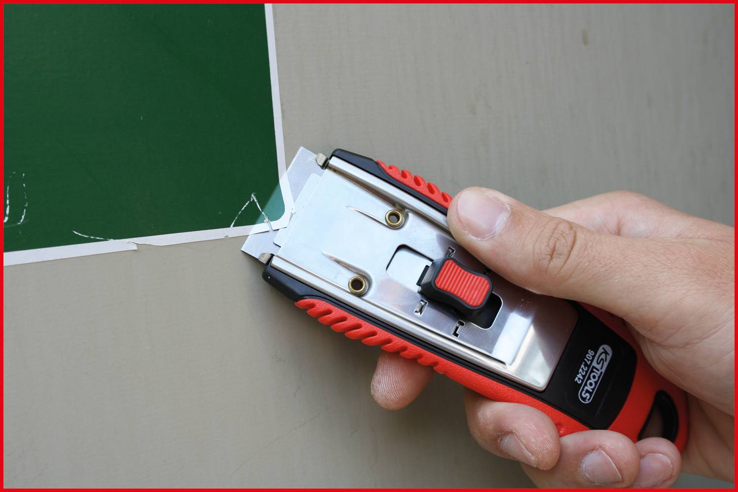 Dłoń trzyma czerwony nóż do tapet z czarnym uchwytem i przycina zieloną folię na powierzchni.