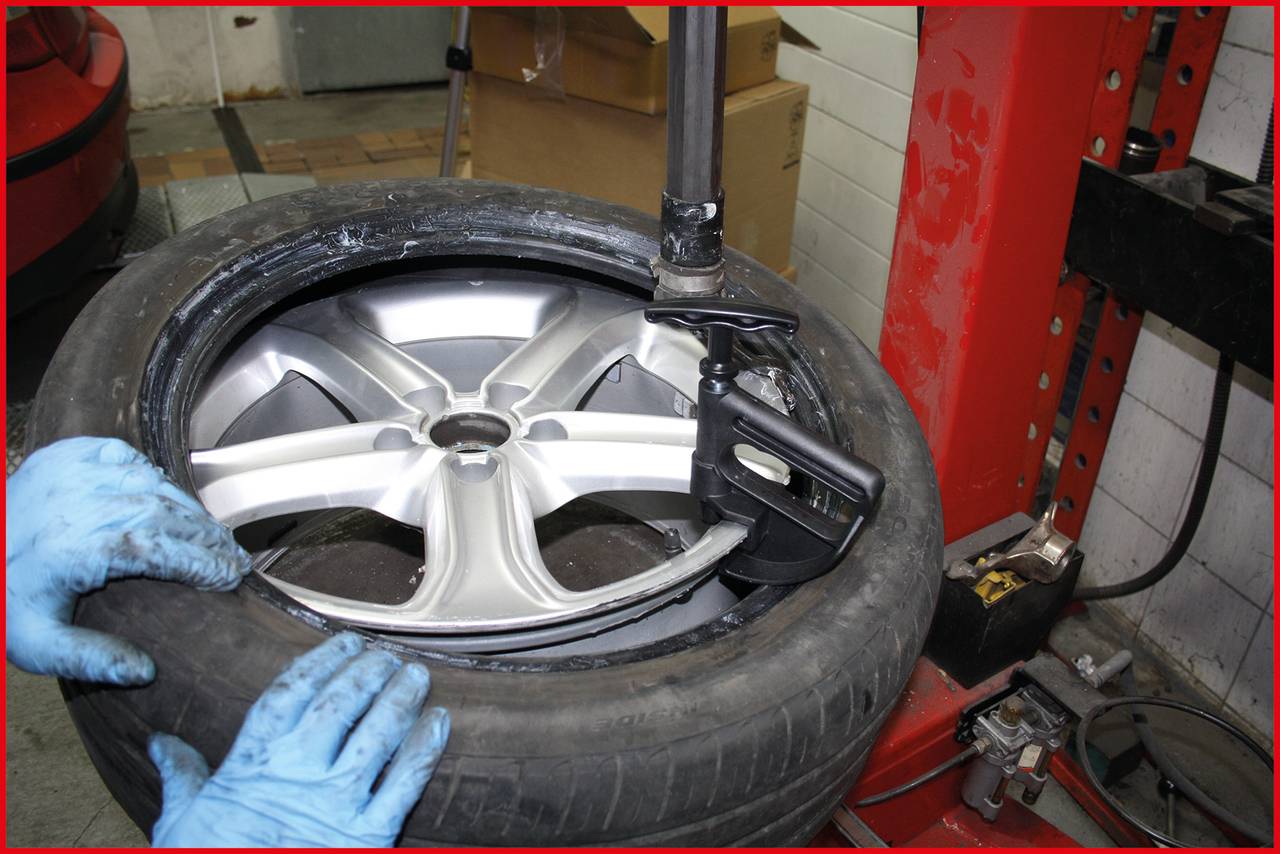 Naprawa opony samochodowej w warsztacie. Dwie ręce w niebieskich rękawiczkach trzymają oponę. Maszyna zdejmuje oponę z felgi.
