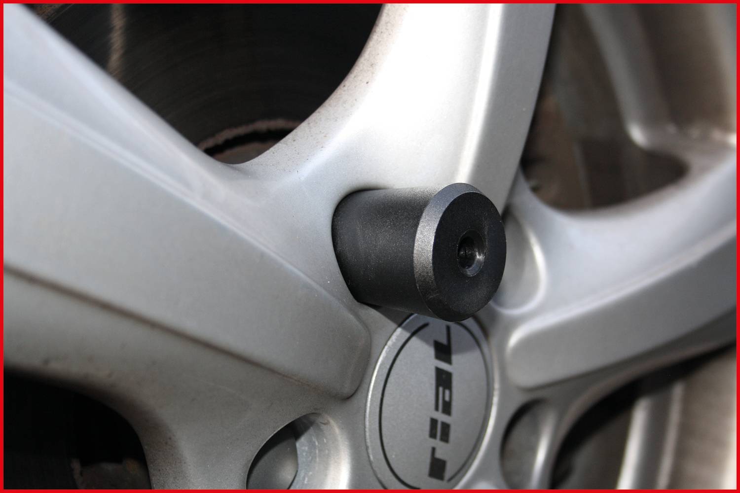 Srebrna felga samochodowa z czarnym, cylindrycznym elementem wyposażenia zamocowanym na śrubie koła.