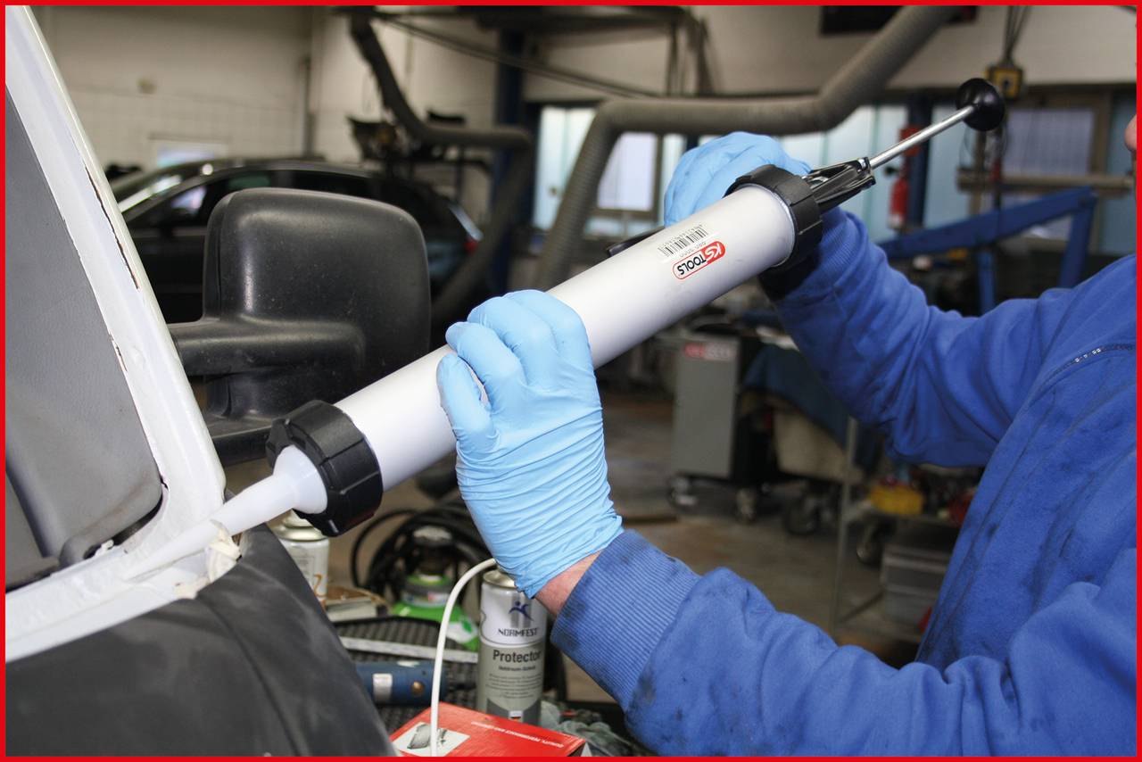 Osoba ubrana w niebieski kombinezon i rękawice, używa białego aplikatora do kleju/uszczelniacza na styku karoserii samochodu w warunkach warsztatowych.