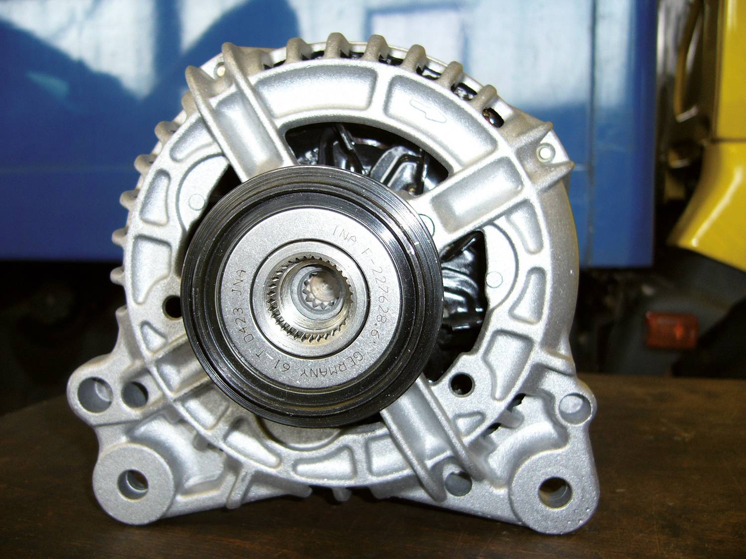 Srebrzysta alternator leżący na stole, z dużymi metalowymi cewkami i centralnym kołem pasowym, na niebieskim tle.