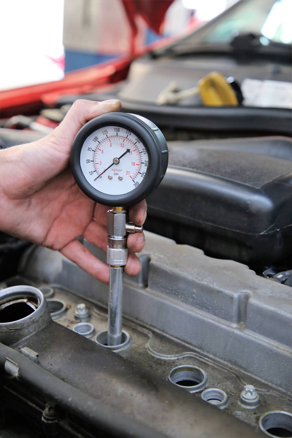 Ręka trzyma manometr podłączony do silnika samochodowego, mierzący ciśnienie. W tle widać komorę silnikową.