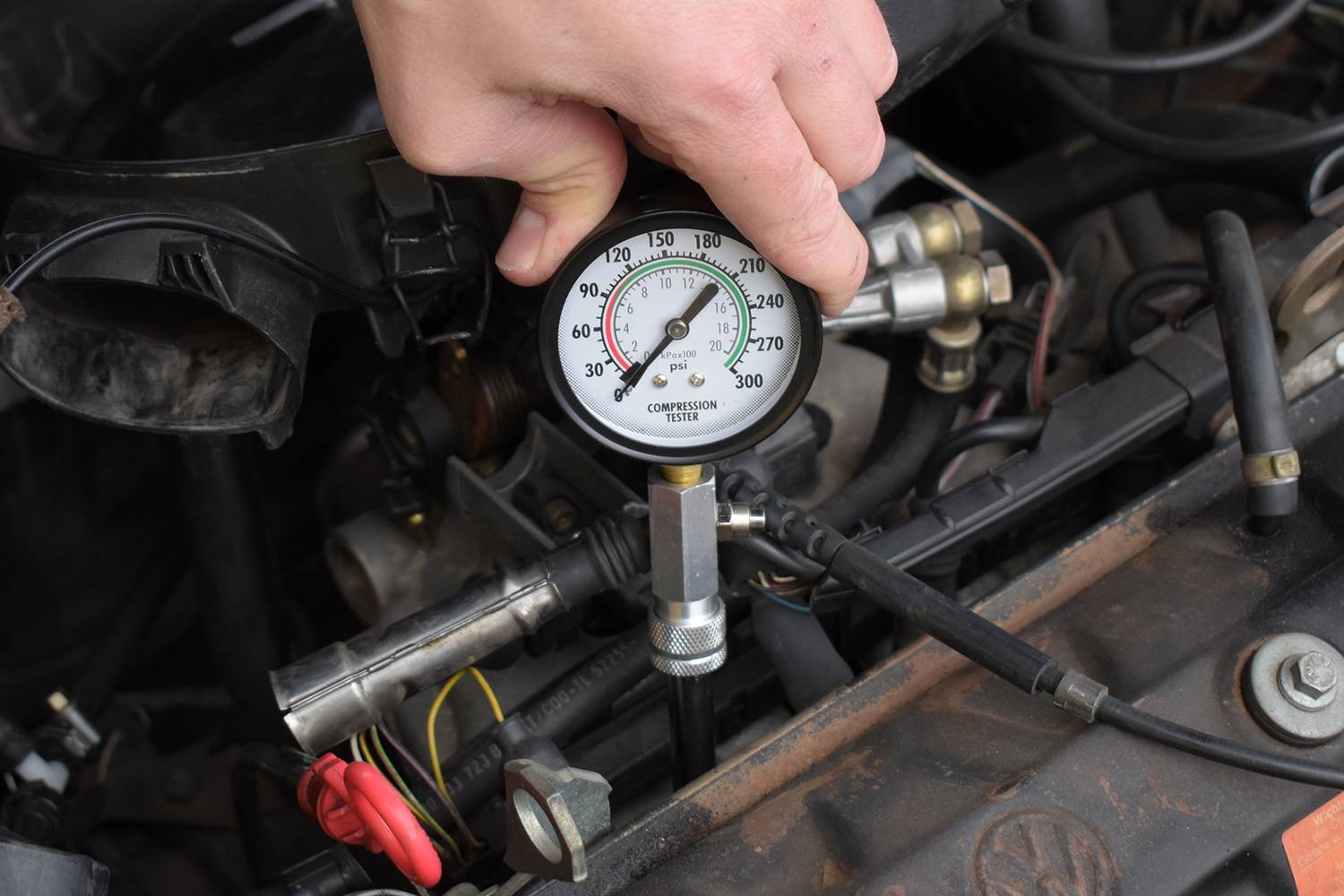 Jedna ręka trzyma urządzenie do pomiaru kompresji nad odkrytym silnikiem. Urządzenie wskazuje około 210 PSI. W tle widoczne są części silnika.