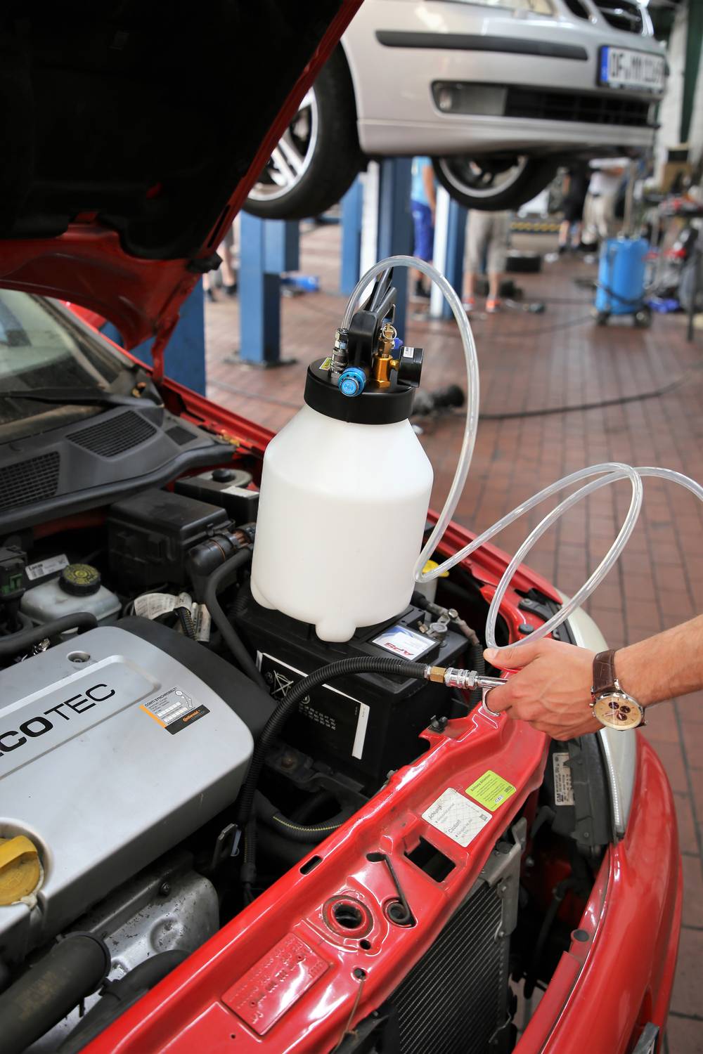 Warsztat przedstawia osobę wlewającą olej do samochodu. Pojemnik z wężem jest podłączony do silnika. W tle widać kolejne samochody na podnośnikach.