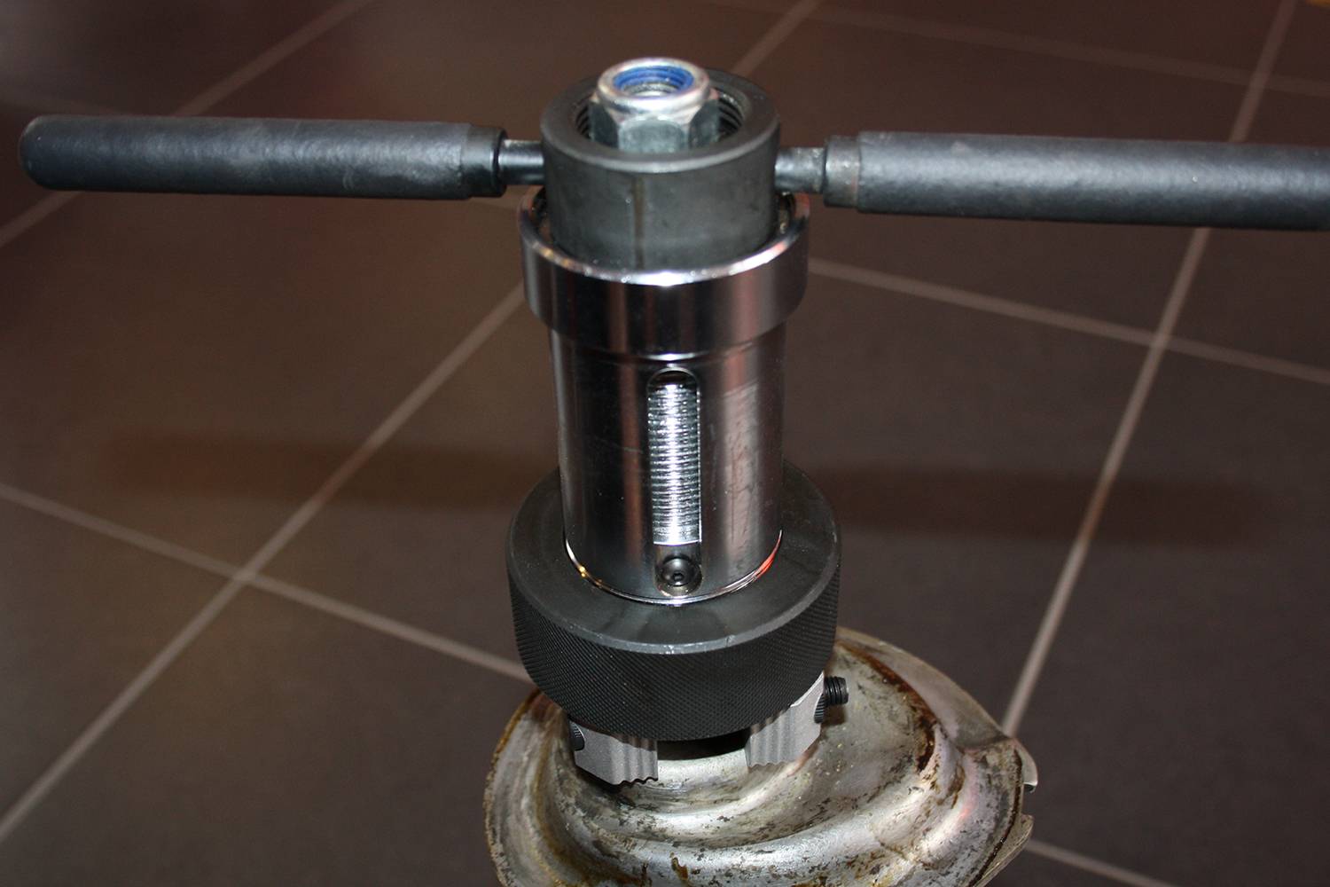 Zbliżenie metalowego narzędzia z uchwytem i gwintem, zamontowanego na statywie, na tle płytek ceramicznych.
