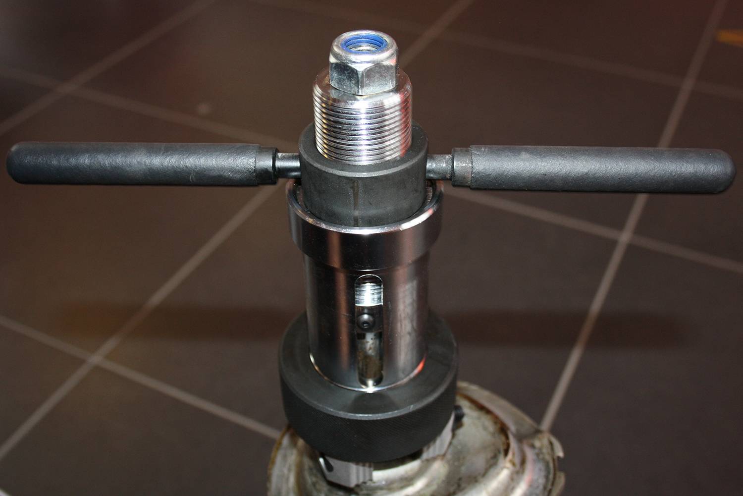 Metalowe narzędzie z dwoma uchwytami, stojące na podłodze wyłożonej płytkami. Wygląda na ręczne urządzenie do obróbki rur.