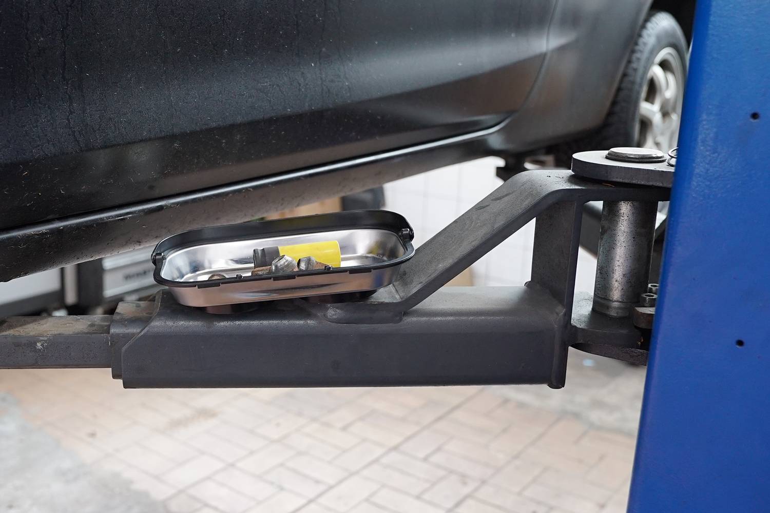 Czarny samochód stoi na podnośniku w warsztacie. Metalowa miska z śrubami i narzędziami leży na wsporniku podnośnika.