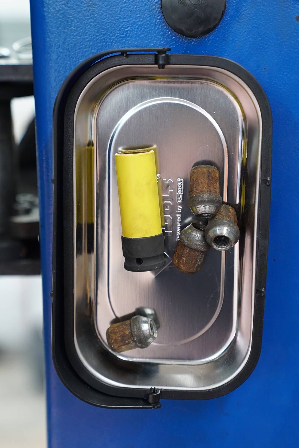 Metalowy pojemnik przy niebieskiej maszynie zawiera żółty adapter oraz kilka zardzewiałych nakrętek.