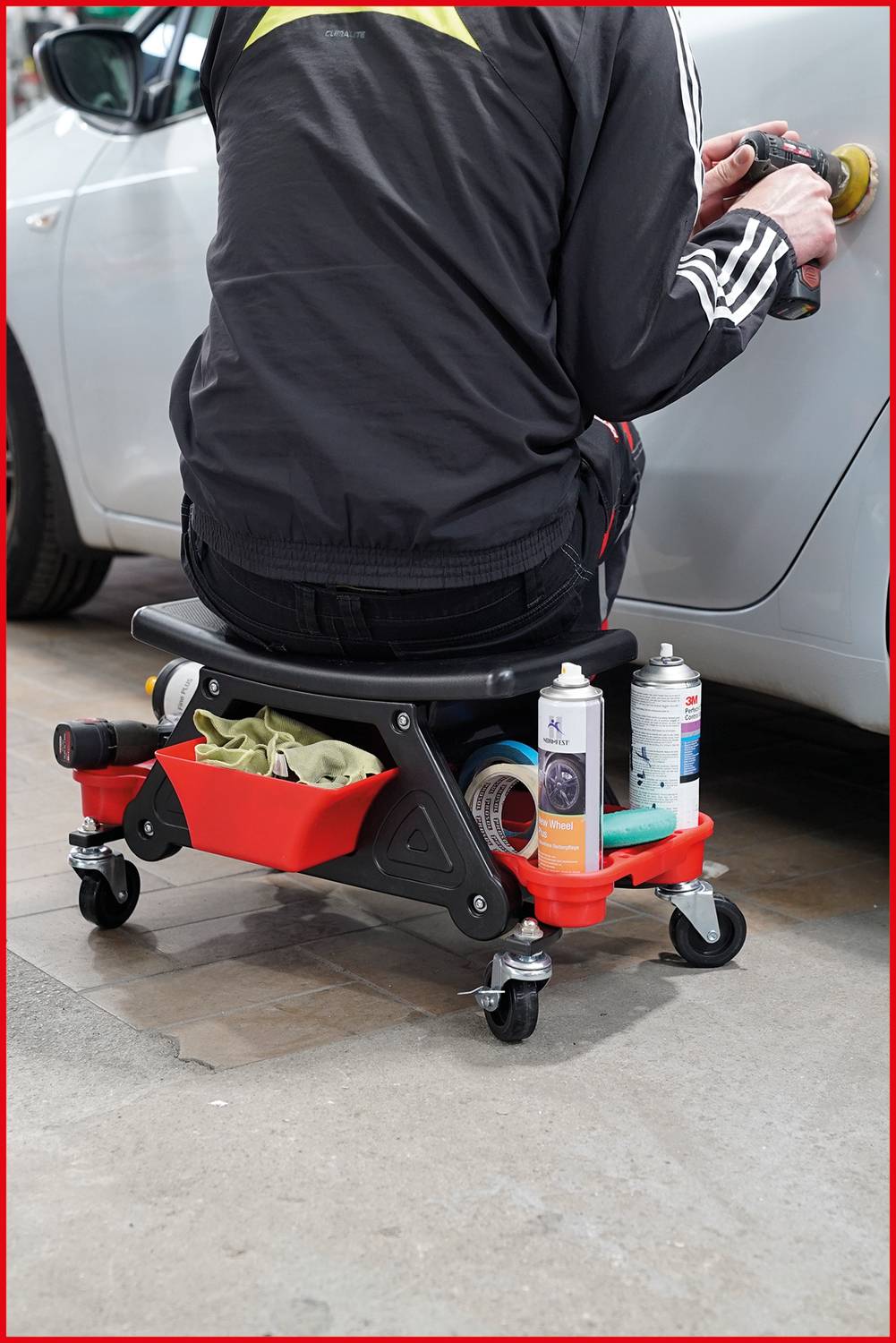 Osoba siedzi na mobilnym stołku warsztatowym i poleruje samochód, podczas gdy narzędzia i produkty pielęgnacyjne są widoczne na półce.