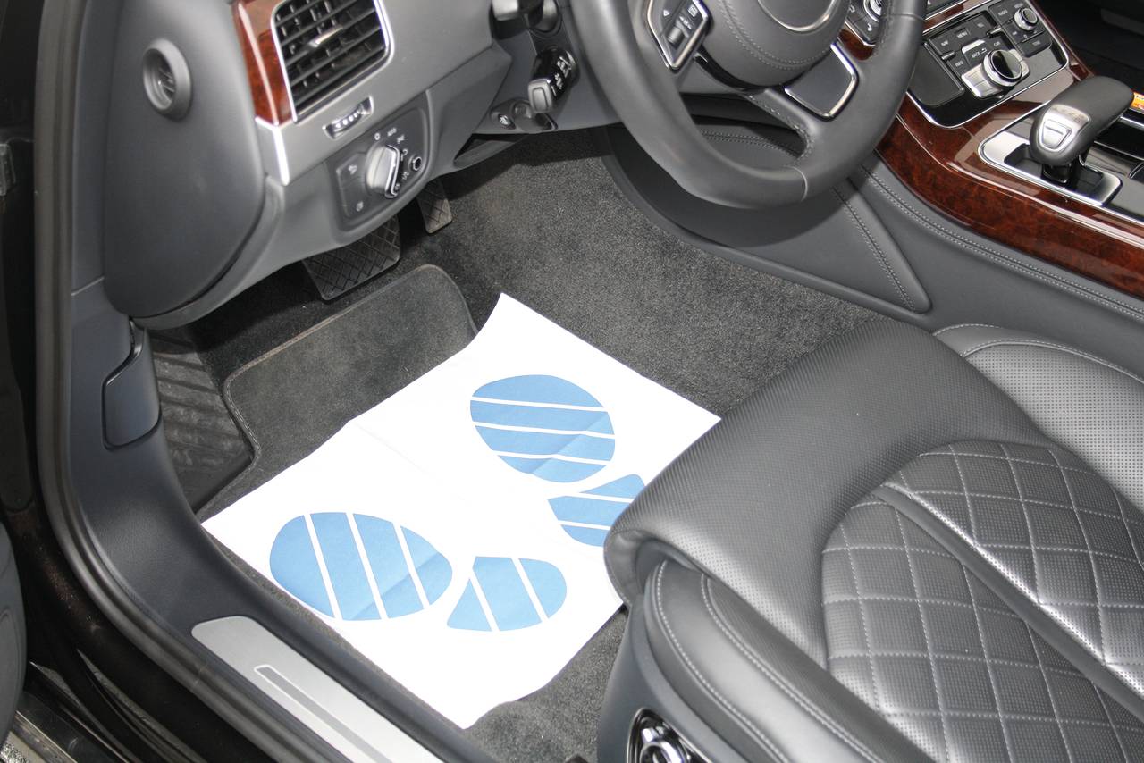 Wnętrze pojazdu z kartką papieru na podłodze, na której widnieją dwa niebieskie ślady stóp. Widoczne kierownica i elementy sterujące.