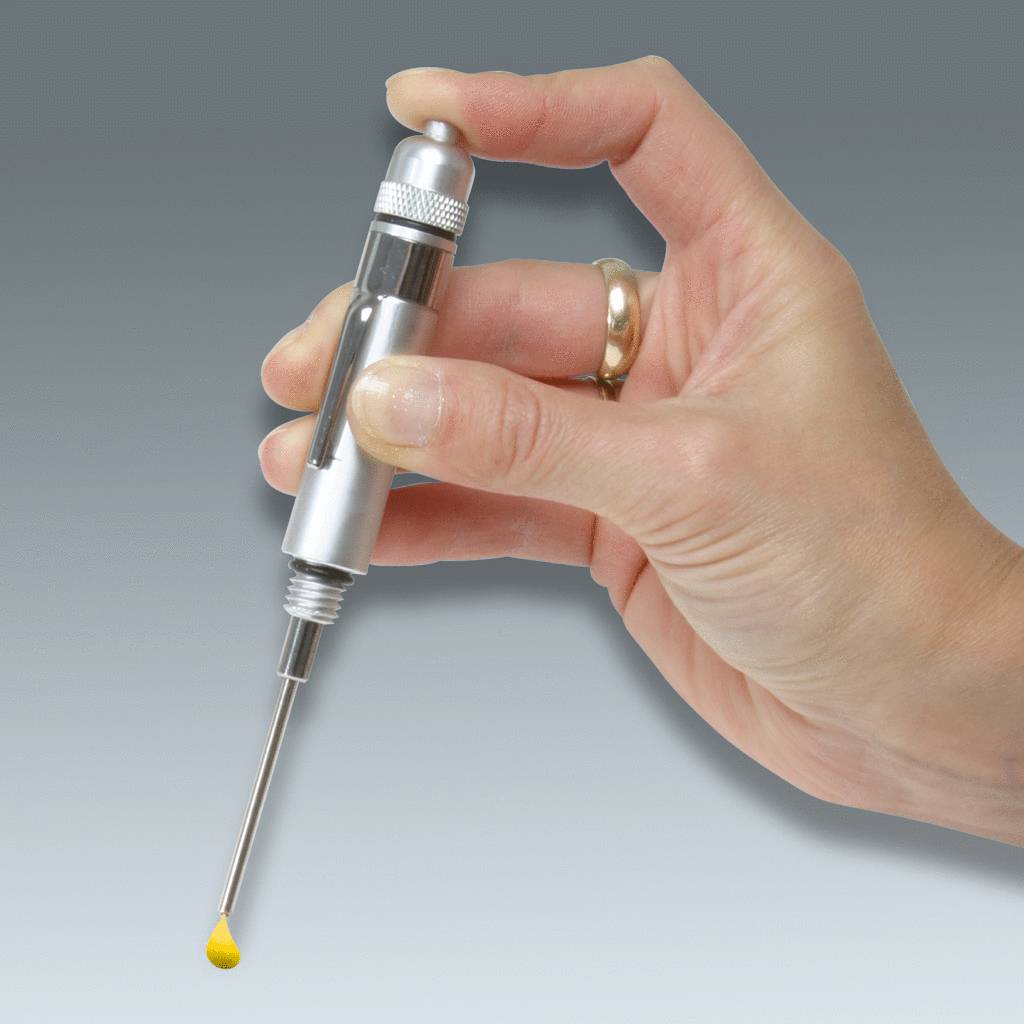 Dłoń trzyma srebrną pipetę z żółtą kroplą płynu na czubku. Tło jest szare.