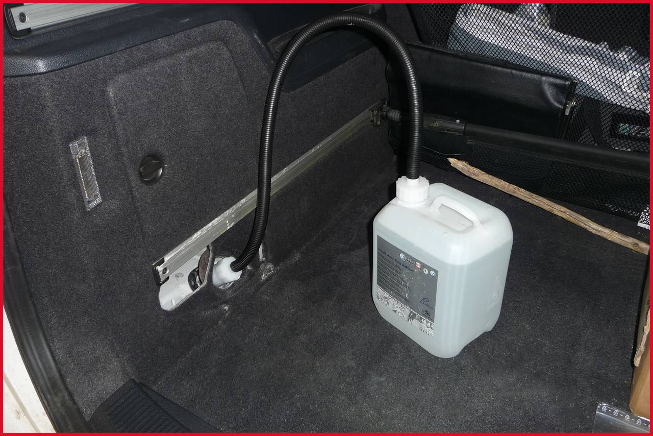 Biały kanister z przymocowaną rurką znajduje się w bagażniku pojazdu. Kanister wydaje się zawierać specjalną ciecz.