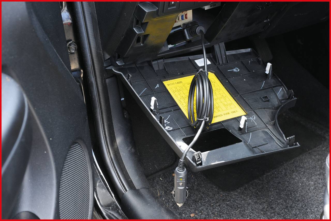 Otwarta schowka w samochodzie z wyciętym dnem, widoczny żółty znak ostrzegawczy, przewód zwisający.