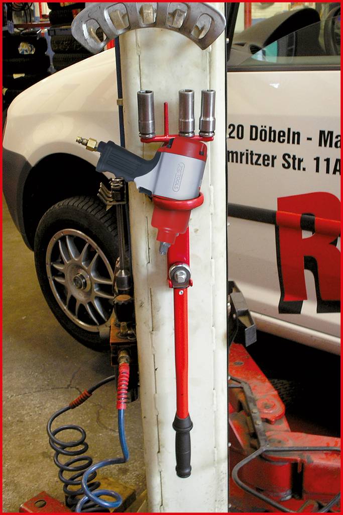 Pneumatyczny klucz do nakrętek kół wisi na stojaku warsztatowym. W tle widać białe auto.