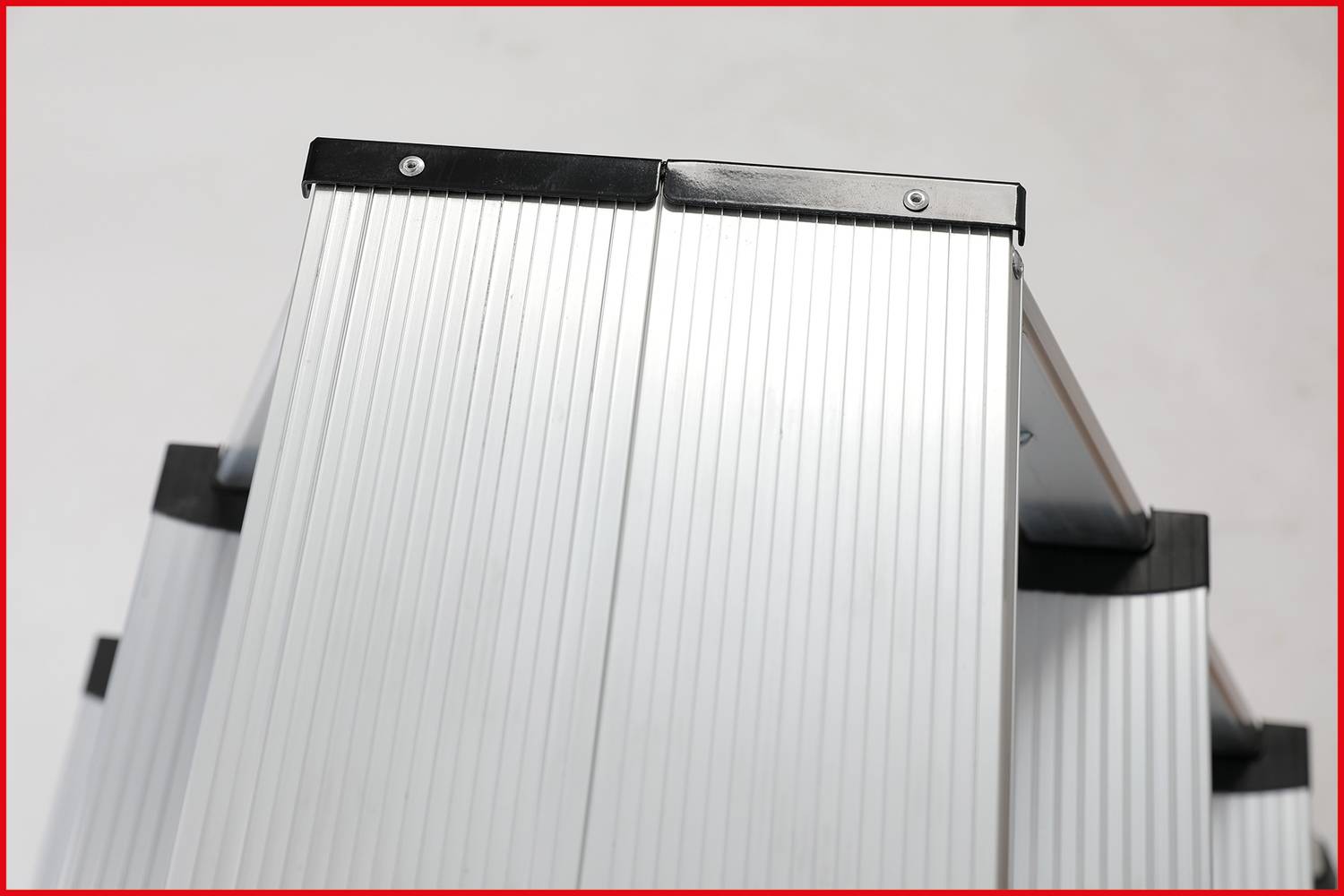 Srebrzysta aluminiowa drabina sfotografowana z przodu. Stopnie są żebrowane dla lepszej przyczepności. Górna część drabiny na neutralnym tle.