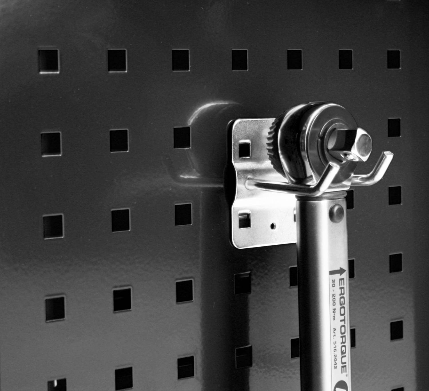 Klucz dynamometryczny wisi na ściance perforowanej. Klucz jest metaliczny i błyszczący, obok niego znajdują się symetryczne otwory.