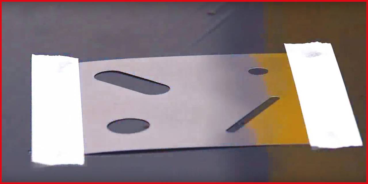 Cienka, prostokątna blacha metalowa z różnymi owalnymi i prostymi wycięciami, zamocowana na ciemnym podłożu.