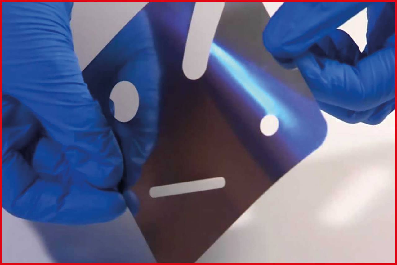 Dwie dłonie w niebieskich rękawiczkach trzymają metalową folię odblaskową z wyciętymi kształtami twarzy na białym tle.