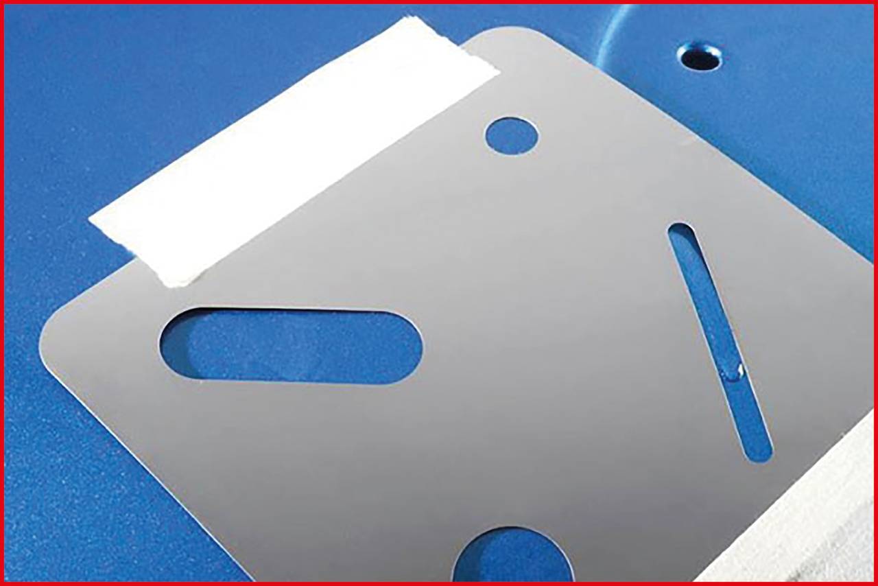 Lśniąca, srebrzysta płyta metalowa z kilkoma wycięciami spoczywa na niebieskiej powierzchni. Na górze przyklejony jest kawałek białej taśmy.