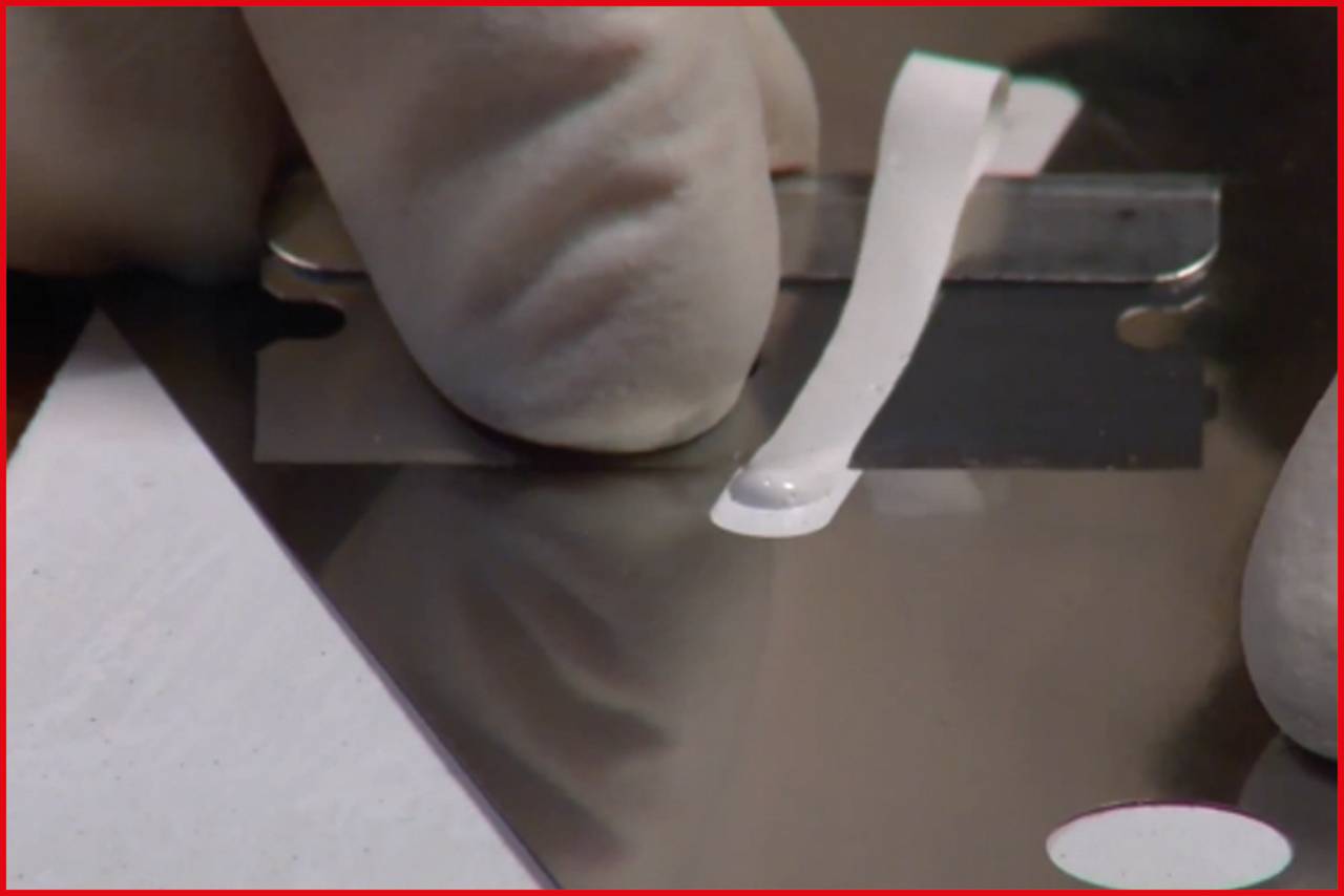 Palec w rękawicy zeskrobuje białą pastę z gładkiej metalowej płyty przy pomocy żyletki, prawdopodobnie w ramach procedury pobierania lub obróbki materiału.