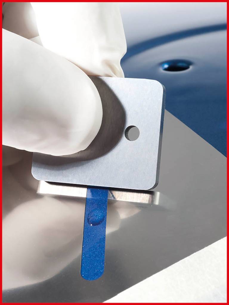 Dłoń w rękawiczce trzyma metalową płytkę z małym otworem, podczas gdy pod spodem niebieski krople spoczywa na lustrzanej powierzchni.