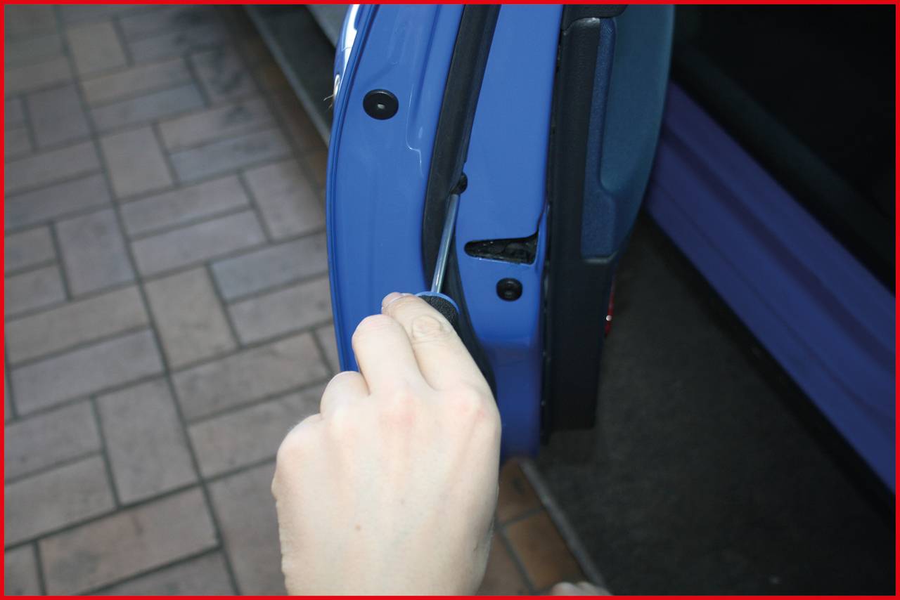 Ręka otwiera drzwi samochodu płaskim narzędziem, prawdopodobnie w celu naprawy lub odblokowania. W tle widoczne są kamienne krawężniki.