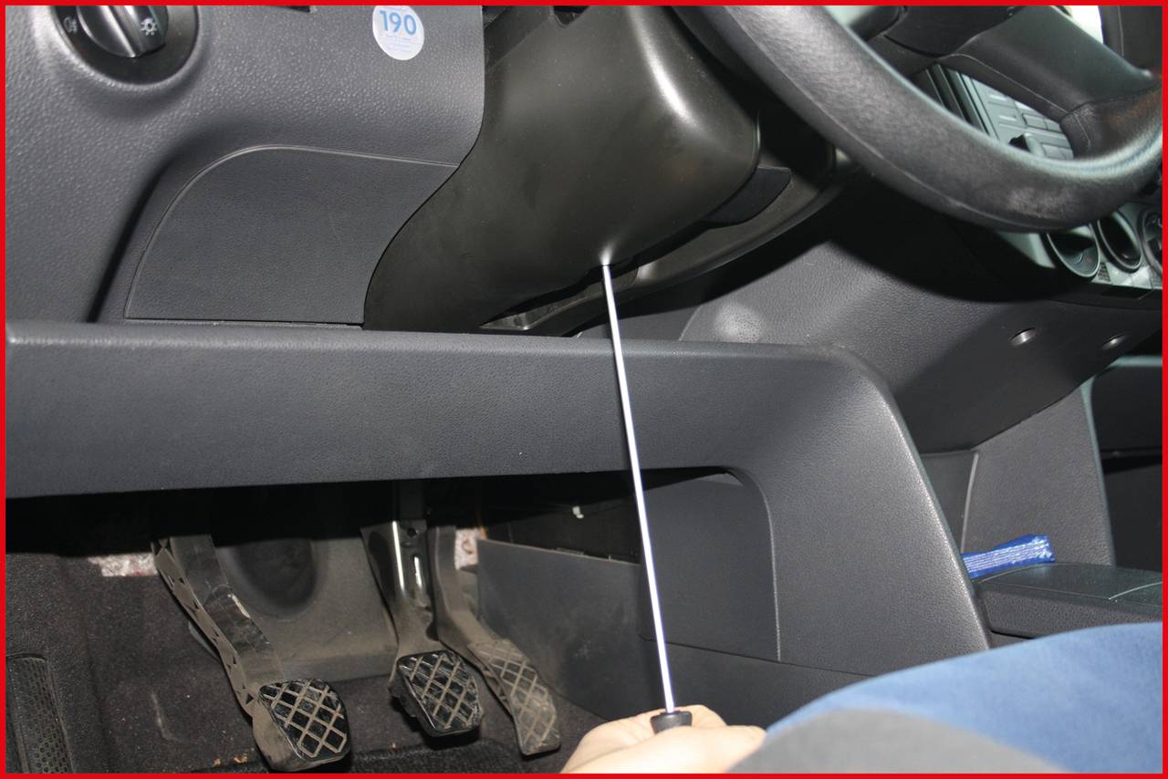 Jedna ręka trzyma śrubokręt wprowadzany pod kierownicą samochodu. Na pierwszym planie widoczne są pedały.