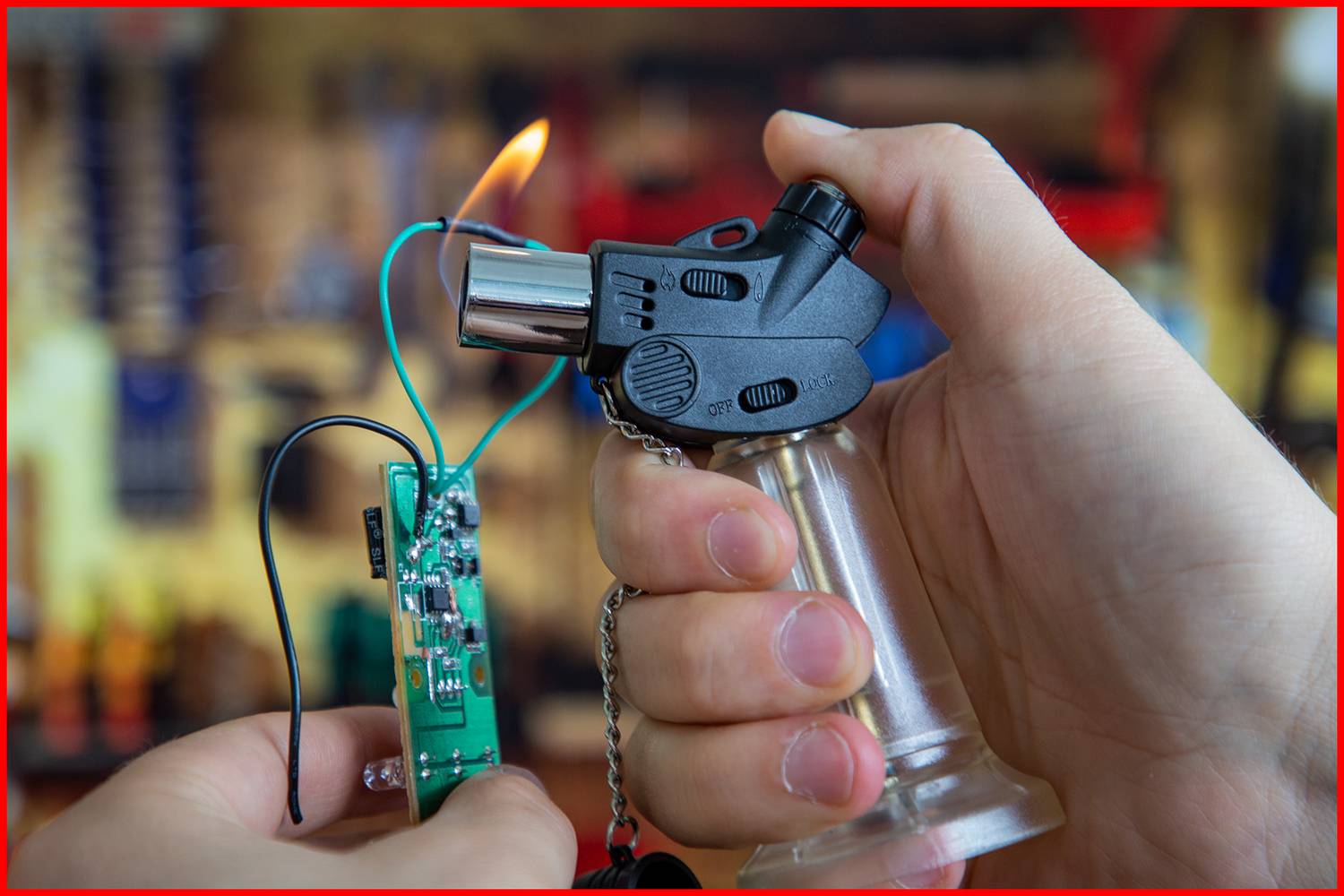 Dłoń trzyma zapalniczkę, która nagrzewa drut z małym obwodem elektronicznym. W tle nieostry, prawdopodobnie środowisko warsztatu.