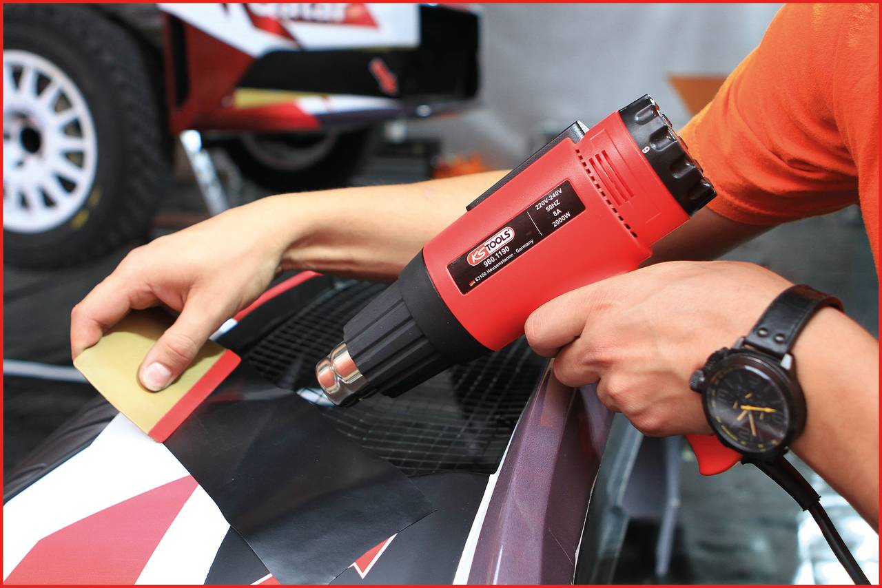 Osoba używa pistoletu na gorące powietrze do przymocowania folii na części samochodowej. W tle widoczne są opony samochodowe.