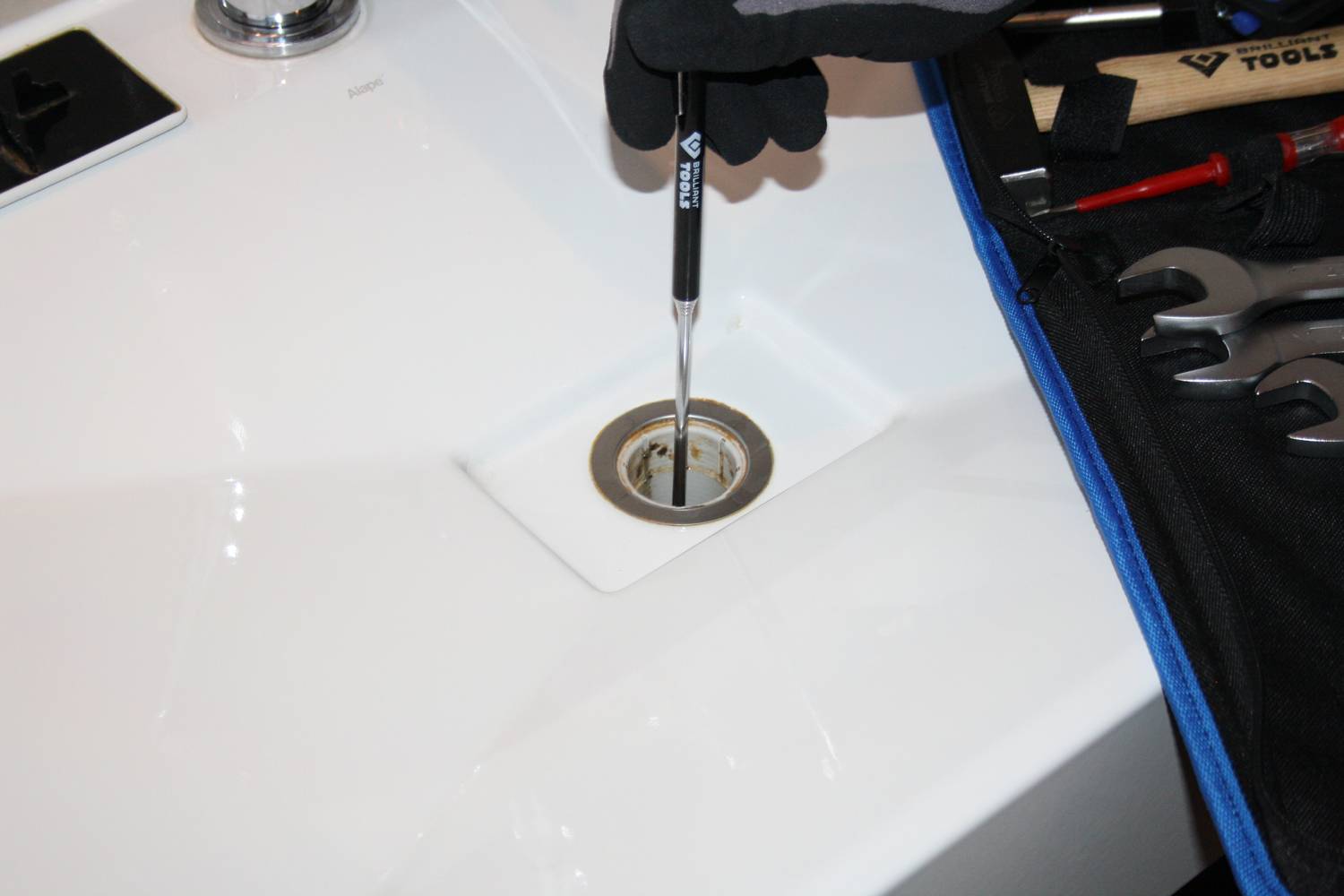 Osoba pracuje narzędziem przy odpływie zlewu. Kilka narzędzi leży na płóciennej torbie obok zlewu.